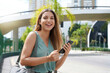 © zigres - Half length portrait of cheerful Brazilian female on street using smartphone. Beautiful millennial girl looking at camera in the city holding mobile phone.
