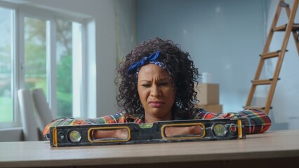 A young African American woman poses next to a building bubble level on a table. The black woman looks at the level in confusion. The concept of repair, finishing work, interior design.