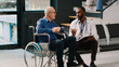 © DC Studio - African american doctor doing consultation with old man in wheelchair, filling in medical report papers at hospital lobby. Patient with chronic disability in waiting room chatting with physician.