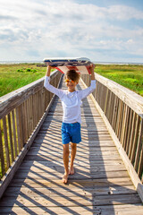 Wall Mural - Young Preteen Boy Wearing Sunglasses and Walking with Boogie Board Over His Head On A Boardwalk