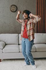 Sticker - Full body fun young Indian man wear casual clothes raise up hands listen music in headphones stand near grey sofa couch stay at home hotel flat rest relax spend free spare time in living room indoor.