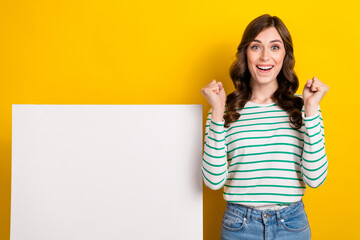 Canvas Print - Photo of impressed ecstatic girl with wavy hairdo dressed striped shirt clenching fist near white board isolated on yellow color background