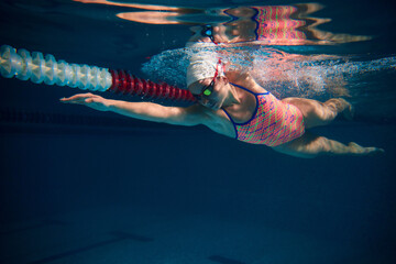 Coordination of movements. Professional female swimmer in cap and goggles in motion, training in swimming pool indoor. Underwater view. Sport, endurance, competition, energy, healthy lifestyle concept