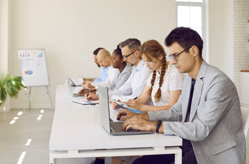 Group of busy colleagues are reviewing documents in preparation for business conference. Side view of men and women with laptops and clipboards sitting in row at long table in bright office.