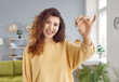 Portrait of a young smiling pretty curly redhead woman looking at the camera with the keys in her hands standing at home. Happy joyful new homeowner enjoing real estate or car purchase.