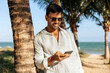© kleberpicui - Smiling man using smartphone on tropical beach under shade of palm trees