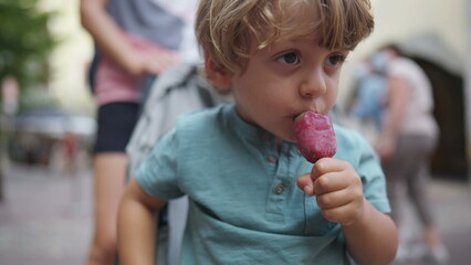 One cute small boy eating ice cream on stick portrait face close up. Kid snacking during summer day outside seated on stroller