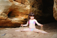 A Little Beautiful Girl Trains Stretching And Plays With Sand On The Beach Near The Cave In Summer.