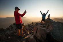 A Hiker Uses A Tablet To Take A Picture Of His Friend Standing Next To The Summit Cairn Of Sauk Mountain.