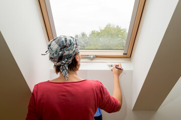 Wall Mural - Woman paints a window recess of a roof window with a brush - painting concept