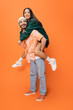 full length of excited man in beanie and jeans piggybacking trendy asian woman on orange background.