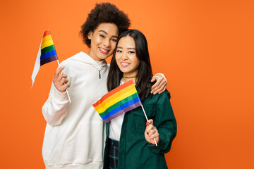 happy multiethnic women in trendy clothes holding lgbt flags isolated on orange.