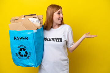 Wall Mural - Young redhead woman holding a recycling bag full of paper to recycle isolated on yellow background with surprise expression while looking side