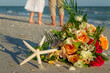Bridal bouquet and starfish with wedding rings on sandy beach with waist down of bride and groom walking away in background