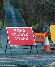 Road Closed Ahead Sign At A Barrier With Traffic Cone And Road Disappearing Into The Distance
