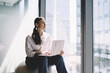 © BullRun - Thoughtful woman sitting with laptop near glass wall