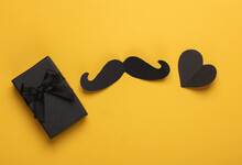 Black Paper Mustache With A Heart And Gift Box On Yellow Background. Father's Day. Top View. Flat Lay