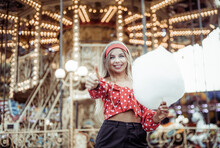 Adorable Cute Beautiful Woman With Cotton Candy Stands In The Middle Of An Amusement Park With Bright Colors, Positive And Cheerful, Happy And Optimistic Emotion