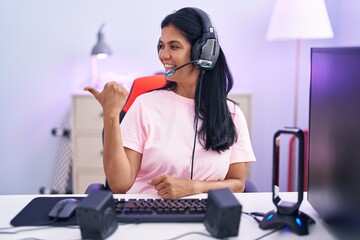 Poster - Mature hispanic woman playing video games at home smiling with happy face looking and pointing to the side with thumb up.