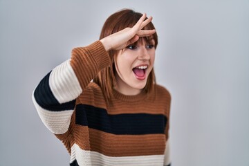 Wall Mural - Young beautiful woman wearing striped sweater over isolated background very happy and smiling looking far away with hand over head. searching concept.