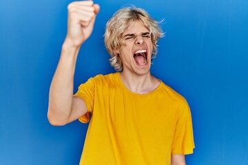 Poster - Young modern man standing over blue background angry and mad raising fist frustrated and furious while shouting with anger. rage and aggressive concept.