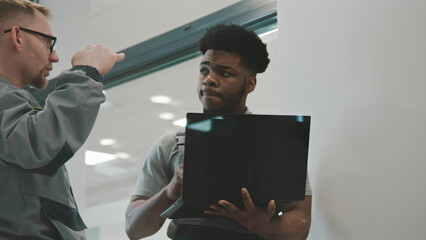Two multi ethnic men in uniform discuss installation of CCTV cameras in office room. Installers check and set up security cameras using computer software on laptop. Concept of surveillance system.