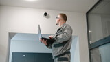 Fototapeta  - Installer in uniform sets up security camera in office room using laptop. Man in glasses checks CCTV cameras in computer program. Monitoring system. Concept of surveillance system and privacy.