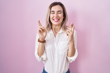 Canvas Print - Young beautiful woman standing over pink background gesturing finger crossed smiling with hope and eyes closed. luck and superstitious concept.