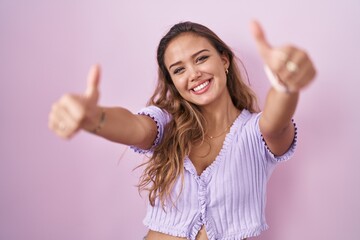 Sticker - Young hispanic woman standing over pink background approving doing positive gesture with hand, thumbs up smiling and happy for success. winner gesture.