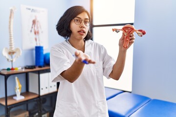 Poster - Young latin woman wearing physiotherapist uniform holding anatomical model of uterus at physiotherapy clinic