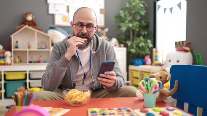 Sticker - Young bald man preschool teacher using smartphone eating chips potatoes at kindergarten