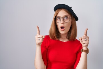 Wall Mural - Young redhead woman standing wearing glasses and beret amazed and surprised looking up and pointing with fingers and raised arms.