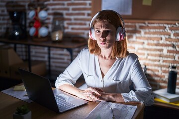 Wall Mural - Young redhead woman working at the office at night wearing headphones puffing cheeks with funny face. mouth inflated with air, catching air.