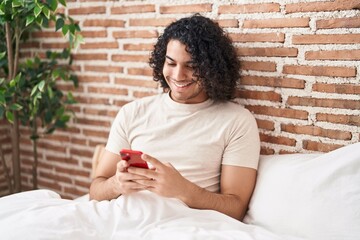 Sticker - Hispanic man with curly hair using smartphone sitting on the bed looking positive and happy standing and smiling with a confident smile showing teeth