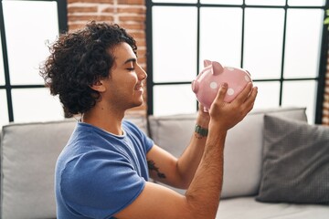 Sticker - Young hispanic man holding piggy bank sitting on sofa at home