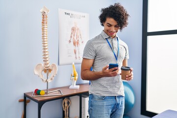 Sticker - Young hispanic man physiotherapist using smartphone and drinking coffee at rehab clinic