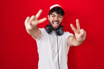 Wall Mural - Hispanic man with beard wearing gamer hat and headphones smiling with tongue out showing fingers of both hands doing victory sign. number two.