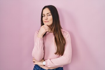 Poster - Young brunette woman standing over pink background looking confident at the camera smiling with crossed arms and hand raised on chin. thinking positive.