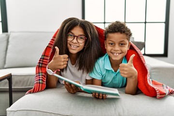 Canvas Print - Two siblings lying on the sofa reading a book doing happy thumbs up gesture with hand. approving expression looking at the camera showing success.