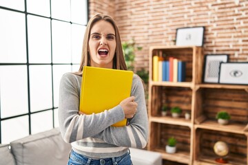 Canvas Print - Young woman holding notebook angry and mad screaming frustrated and furious, shouting with anger. rage and aggressive concept.