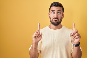 Canvas Print - Handsome hispanic man standing over yellow background pointing up looking sad and upset, indicating direction with fingers, unhappy and depressed.