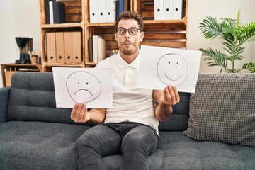 Poster - Young hispanic man working on depression holding sad to happy emotion paper making fish face with mouth and squinting eyes, crazy and comical.