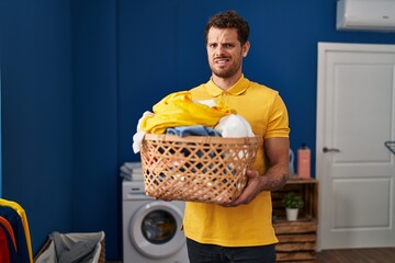 Wall Mural - Young hispanic man holding laundry basket clueless and confused expression. doubt concept.