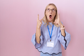 Sticker - Young caucasian business woman wearing id card amazed and surprised looking up and pointing with fingers and raised arms.