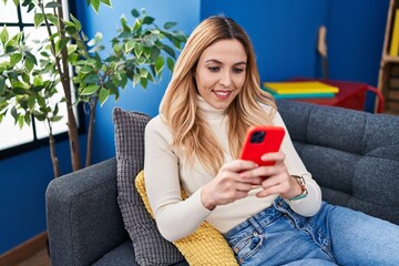 Wall Mural - Young blonde woman using smartphone sitting on sofa at home