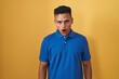 Young hispanic man standing over yellow background in shock face, looking skeptical and sarcastic, surprised with open mouth