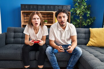 Poster - Young couple playing video game holding controller at home scared and amazed with open mouth for surprise, disbelief face