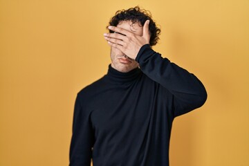 Wall Mural - Hispanic man standing over yellow background covering eyes with hand, looking serious and sad. sightless, hiding and rejection concept