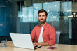 © Liubomir - Portrait of a smiling and successful mature man in the office, a businessman with a beard and a red shirt is smiling and looking at the camera, a programmer is using a laptop at work, a creative