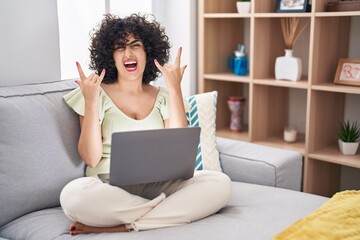 Sticker - Young brunette woman with curly hair using laptop sitting on the sofa at home shouting with crazy expression doing rock symbol with hands up. music star. heavy concept.